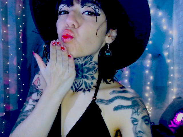 LannieGonzales, con tatuajes de estampados de tigre en tinta negra en el cuello y los brazos, hace un puchero con la mano cerca de la cara, labios rojos, sombrero negro. Superficie negra, luces de colores azul y blanco en el fondo. Increíblemente atractivo/a..