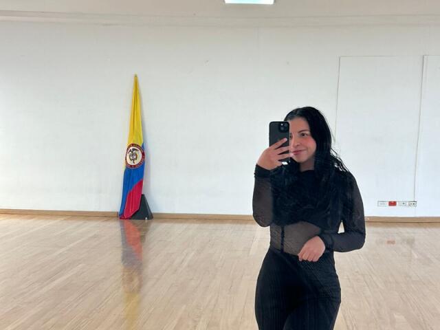 IanaPrince staat in een helder verlichte kamer en maakt een selfie met een zwarte telefoon. Ze draagt een doorschijnend zwart shirt met lange mouwen, met de Venezolaanse vlag op de achtergrond. Houten vloer, witte muren
.