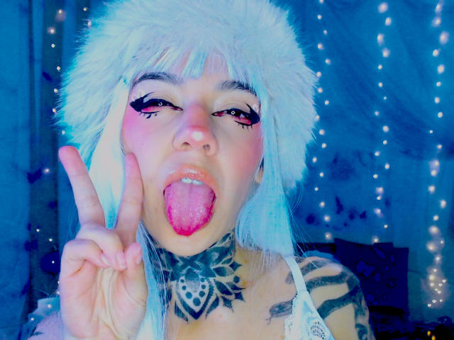 Nahaufnahme von LannieGonzales mit einem weißen Fellhut, die die Zunge herausstreckt und mit der rechten Hand ein Friedenszeichen macht, blau und rosa Eyeliner, schwarze Stammes-Tattoos, Hintergrund mit leuchtenden blauen Lichterketten..