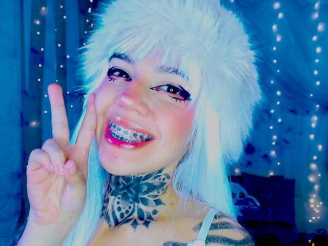 Photo en gros plan de LannieGonzales, faisant un signe de la paix avec un large sourire radieux. Portant un chapeau blanc en fourrure, un collier léopard noir et blanc, et un maquillage bleu éclatant. Fond avec des lumières bleues.