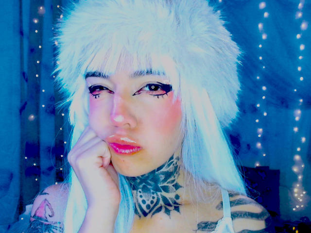 Nahaufnahme von LannieGonzales mit einem hellweißen Fellhut, blauem Leuchten und rosafarbenen Lippen. Sie beißt sich auf den Finger, trägt eine schwarze, gemusterte Halskette und hat neonpinken Lidschatten. Blaue Lichterketten im Hintergrund.