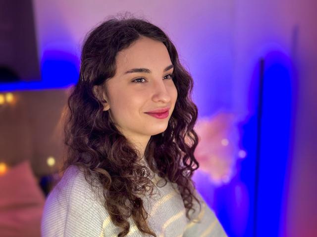 Photograph of JosephineRowe, a curvy woman with long, wavy black hair, wearing a white sweater, smiling slightly, blue and purple lights in the background, glowing softly. Fucking hot.