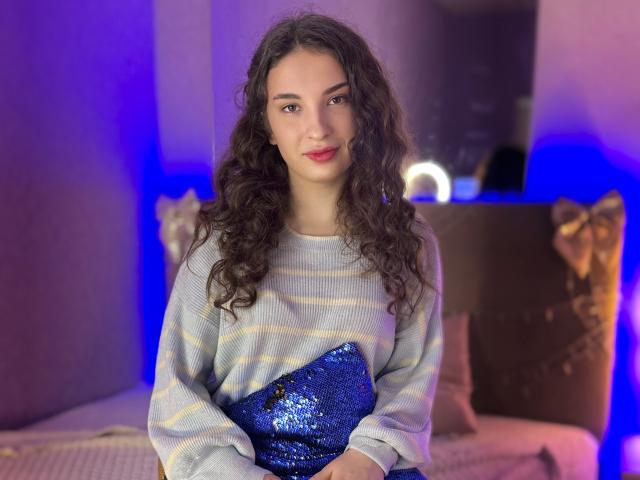 Photograph of JosephineRowe, a curvy woman with long, curly black hair, wearing a white sweater and holding a blue sequined clutch. Soft blue lighting, bedroom setting, slightly seductive pose.