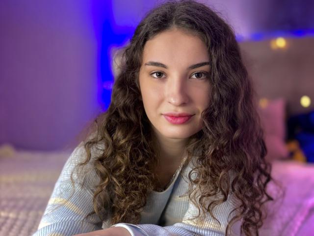 Close-up photograph of JosephineRowe, a young woman with long, curly brown hair, light skin, and red lipstick, leaning forward slightly, wearing a light blue striped shirt, blue and purple lighting.