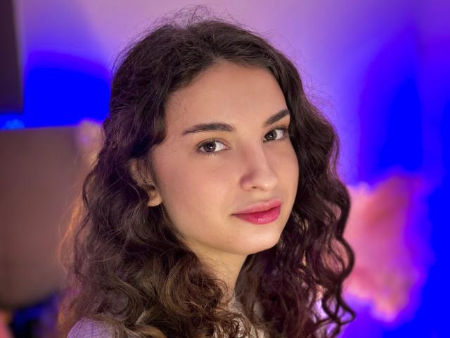 Photograph of JosephineRowe, a young woman with long, wavy black hair, light skin, and red lips, looking directly at the camera. Blue and purple background. Slight sexual tension, fucking gaze.