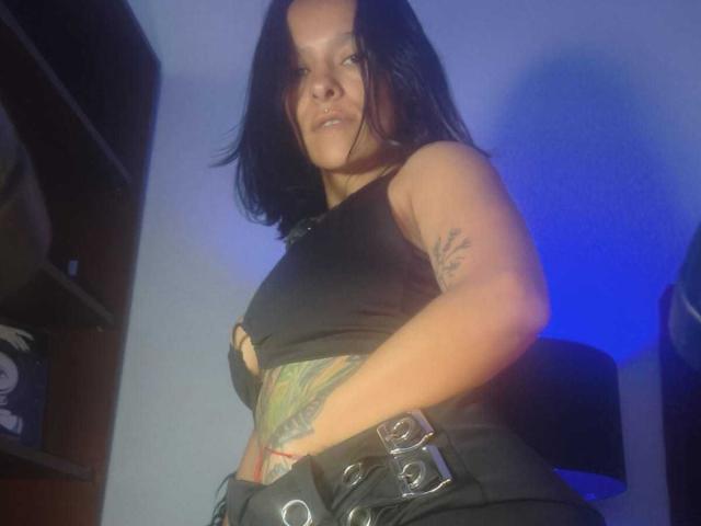 AmelieBlake with shoulder-length black hair, black tank top, and black pants with silver belt loops, stands with hands on hips. Visible tattoo on right side. Blue light background. Fucksack.