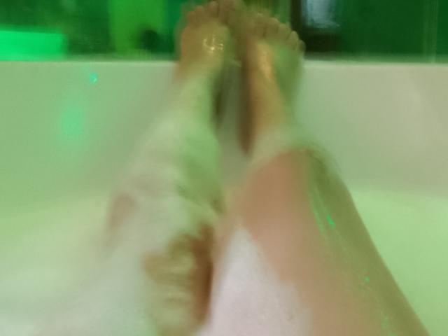 Photo floue des jambes de DruHot dans une baignoire, les pieds se touchant, avec de l'eau verte et des bulles de savon. Des orteils en forme de pénis visibles, une position suggestive, une peau humide, une atmosphère sensuelle. Ambiance légèrement érotique.