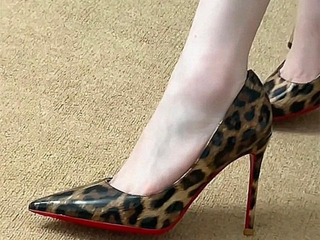 Close-up of Lizee's legs in leopard-print high heels with red soles, standing on a beige carpet. White stockings contrast with the bold pattern and sexy, pointed heels.
