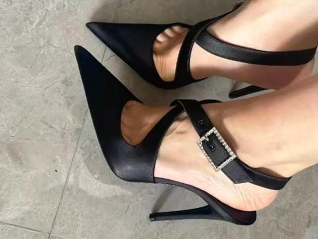 Close-up of Lizee's feet wearing black, pointed high heels with ankle straps and a silver buckle. Her fair skin contrasts with the dark shoes. Sexual tension from the provocative pose.