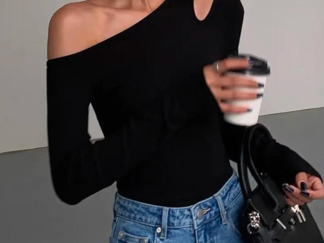 Lizee porte un haut noir à une épaule, tient une tasse de café et est vêtue d'un jean bleu. Sac à main noir avec un pendentif argenté sur le côté droit. Silhouette élancée, légère inclinaison de la tête, style décontracté..
