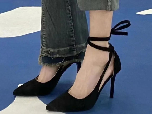 Close-up of Lizee's legs and feet, wearing black high-heeled shoes with ankle straps, and frayed blue jeans that are slightly rolled up. The blue and white background is blurred.