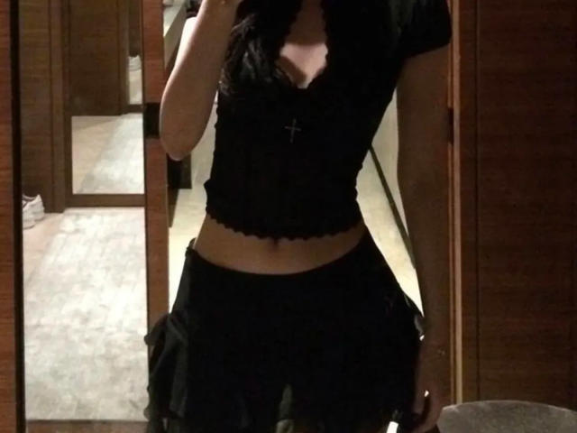 Photograph of a slim person, Lizee, with long dark hair, wearing a black lace-trimmed top and black skirt, standing in a doorway, showcasing a slight tan line on their stomach.