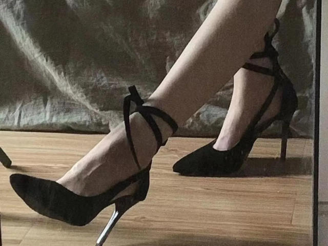 Gros plan des jambes de Lizee, chaussées de talons aiguilles noirs à brides, avec des talons argentés. Ses pieds sont croisés, mettant en valeur ses... peau pâle et le plancher en bois en dessous. Tension sexuelle, jambes, chaussures, pieds croisés.