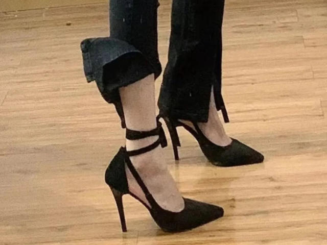 Gros plan des pieds de Lizee, chaussée de chaussures noires à talons hauts avec brides aux chevilles, debout sur un plancher en bois. Le pantalon noir de Lizee est légèrement retroussé, laissant apparaître une peau pâle. Extrêmement séduisant..
