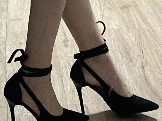 Close-up of Lizee's legs wearing black high-heeled shoes with ankle straps. The shoes are sleek, with pointed toes and thin heels. Shadowed lighting creates a stark contrast. Slight sexual tension.