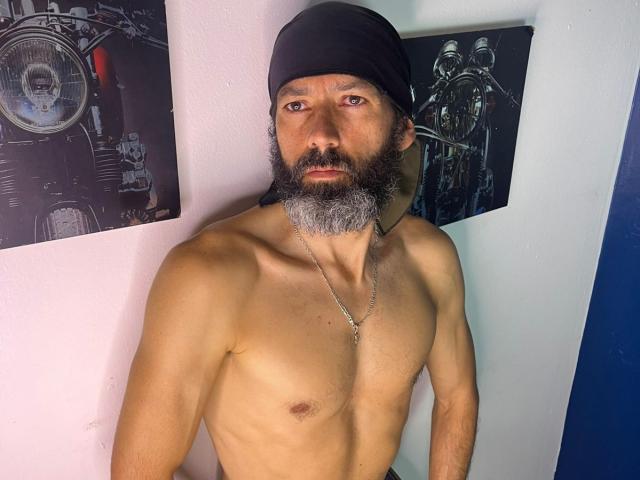 JorgesPerez sans chemise, portant un bonnet noir, une barbe grise et un collier, debout contre un mur avec deux éléments en noir et blanc. Affiches représentant des paysages urbains. Il est musclé, il regarde droit devant lui, avec une érection visible..