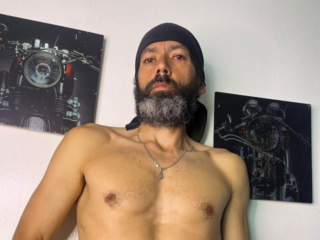 JorgesPerez sem camisa, com barba completa e boné preto, usando um colar de prata, está em frente a uma parede com duas figuras escuras. pinturas abstratas.
seu peito está exposto, mostrando pequenos mamilos..