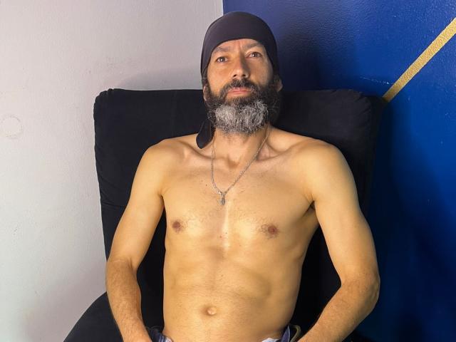 JorgesPerez sin camisa, con un gorro negro, barba gris y collar, sentado en un sofá negro. Sus ojos están medio cerrados, transmitiendo una actitud relajada, ligeramente excitada. Iluminación focalizada.