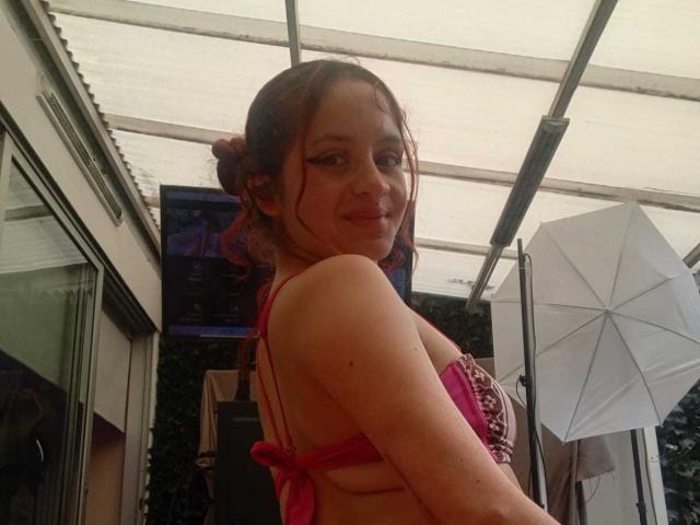 LesleyGray, a young woman with light skin and brown hair in a messy bun, wears a red bikini with pink lace, looking over her shoulder with a playful smile. Sunlit patio, umbrella.