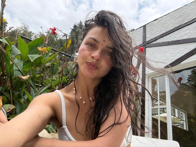 LucyySky, con el cabello oscuro y mojado, se inclina hacia adelante, vistiendo una camiseta blanca que deja ver parte de su escote. Ella está al aire libre, cerca de una estructura de vidrio con plantas y flores rojas en el fondo. Increíblemente atractivo/a..