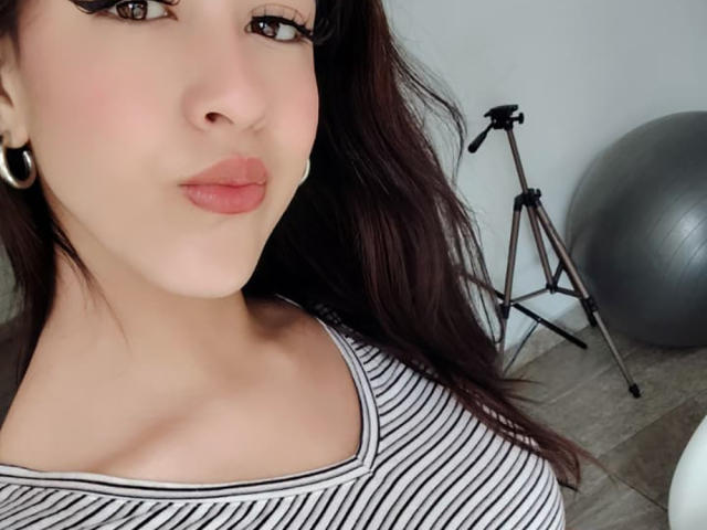 Primer plano de VanellopeLm con piel clara, labios carnosos y cabello castaño oscuro, vistiendo una blusa a rayas, aretes negros y una expresión sugestiva.
un trípode para cámara y una pelota de ejercicio en el fondo..