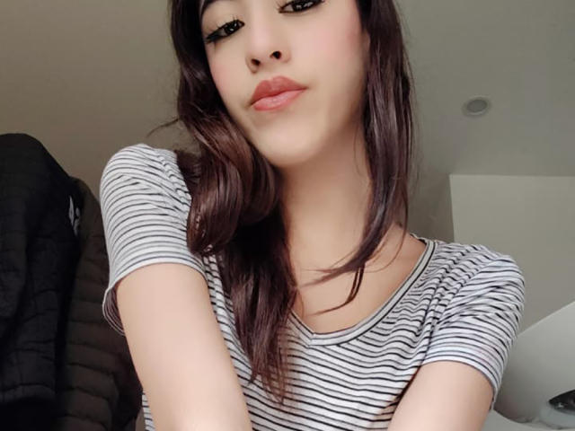 Close-up selfie of VanellopeLm, an Asian woman with long dark hair, wearing a white and black striped shirt. She gazes at the camera with slightly parted lips, conveying subtle sexual tension.