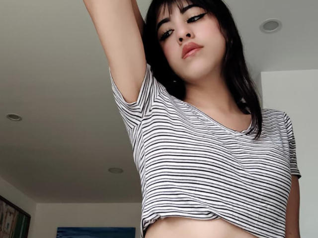 Close-up of VanellopeLm with dark hair, wearing a white and black striped crop top, lifting her arm. She has a slight smirk, pale skin, and black eyeliner. Her chest is visible.
