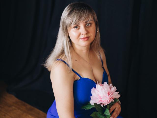 Fotografia de VeronicaSvitt, uma mulher branca com cabelo loiro na altura dos ombros, vestindo um vestido azul justo com um decote baixo. decote, segurando uma flor rosa contra um fundo preto. Ela tem um sorriso discreto..