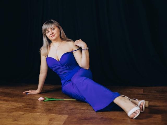 Veronica Svitt está sentada en el suelo de madera, vestida con un vestido azul ajustado y sin tirantes, que deja ver una generosa escote. Se recuesta, con la mano en el muslo, las piernas extendidas, sandalias blancas, pulsera negra, fondo oscuro..
