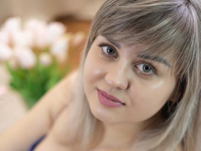 Close-up van VeronicaSvitt, een jonge vrouw met lichtbruin haar en een pony, blauwe ogen en roze lippenstift, die recht in de camera kijkt. naar de camera, met een lichte glimlach. Vervagende witte achtergrond.