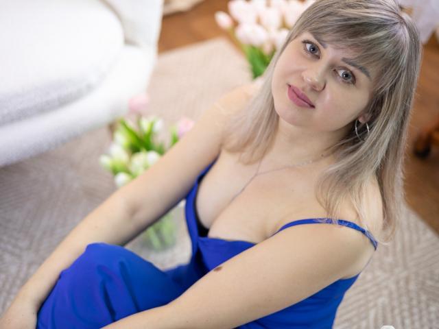 Photograph of VeronicaSvitt, a woman with light brown hair and bangs, wearing a low-cut blue dress, sitting on a carpeted floor. She gazes at the camera with a slight smile. Soft lighting.