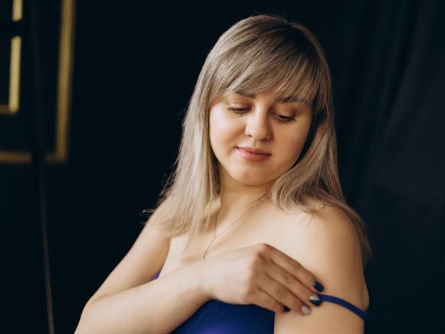 Fotografie von VeronicaSvitt mit hellbraunen Haaren, die nach unten schaut und leicht lächelt, sie trägt ein blaues Top ohne Träger, rechts. Hand, die auf ihrer Brust ruht, weiches Licht betont ihr Gesicht, dunkler Hintergrund..