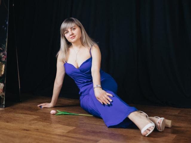 VeronicaSvitt está sentada em um chão de madeira, apoiada em um braço, vestindo um vestido azul justo com um decote baixo, mostrando decote generoso. Ela tem cabelos longos e lisos, sandálias brancas e um laço verde. A expressão dela é sedutora..