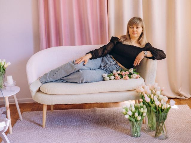 Veronica Svitt è sdraiata su un divano bianco e curvo, con le gambe incrociate, indossando una camicetta di pizzo nero e jeans blu. Fiori rosa e bianchi in vasi di vetro sul pavimento. Luce naturale soffusa. Atmosfera rilassata e sensuale.