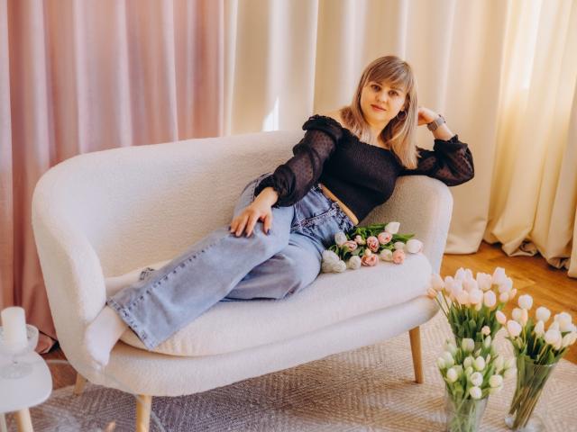 Veronica Svitt zit ontspannen op een witte bank, gekleed in een zwarte doorschijnende top en blauwe jeans, met gekruiste benen en haar hand in haar haar. Bloemenarrangement en witte tulpen op de voorgrond. Zacht licht, ontspannen pose..