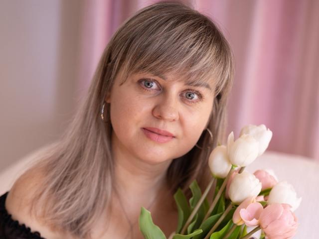 Primer plano de VeronicaSvitt con cabello castaño claro y flequillo, sosteniendo un ramo de tulipanes rosas y blancos. Ella lleva una blusa negra, y en el fondo se ven cortinas rosas suaves. Qué lindo/a..