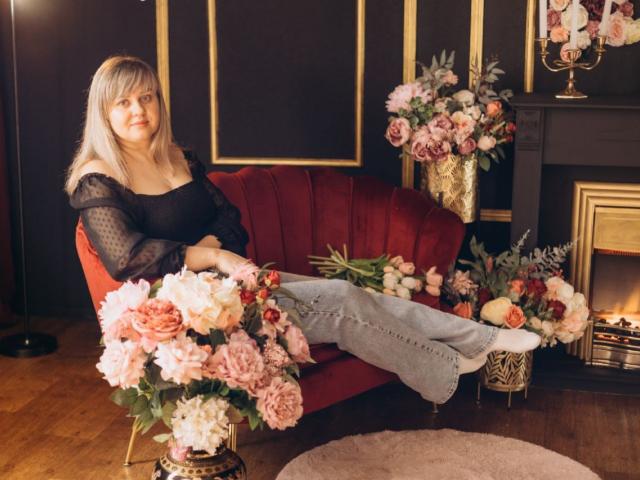 VeronicaSvitt, blond, zwarte top met open schouder, grijze jeans, benen omhoog op een rode bank, omringd door roze en witte rozen. Kachel op de achtergrond, gouden details, zachte verlichting, aantrekkelijk, ontspannen pose..