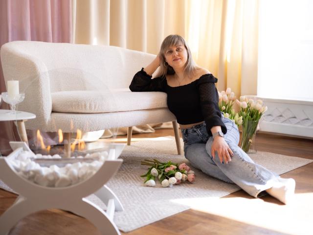 VeronicaSvitt sits on a white rug in a sunlit room, wearing a black off-shoulder top and blue jeans. She leans on a white couch with a flower bouquet nearby. Fucking sexy.