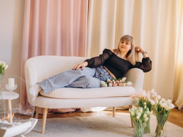 VeronicaSvitt está relaxada em um sofá branco, vestindo uma blusa de malha preta e jeans azuis, com as pernas abertas, a mão no cabelo, e flores por perto. o sofá. Quarto claro, cortinas creme, tulipas brancas. Incrivelmente sexy..