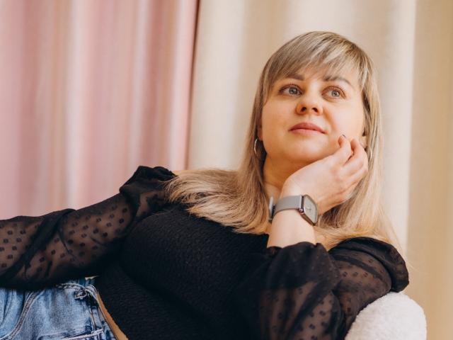 Fotografia de VeronicaSvitt com cabelo loiro na altura dos ombros, olhos azuis e uma blusa preta transparente com bolinhas. Ela está apoiando o queixo na mão, olhando um pouco para o lado, e está usando um relógio cinza..