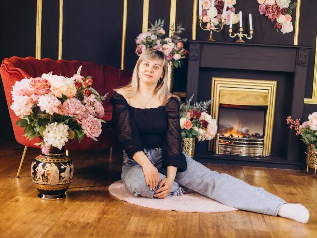Veronica Svitt sitzt auf einem weißen Teppich vor einem brennenden Kamin, trägt eine schwarze Spitzenbluse und graue Hosen, und hat einen Blumenkranz auf. Ein roter Samtstuhl und aufwendig verzierte Vasen mit rosa Blumen. Verdammt heiß..