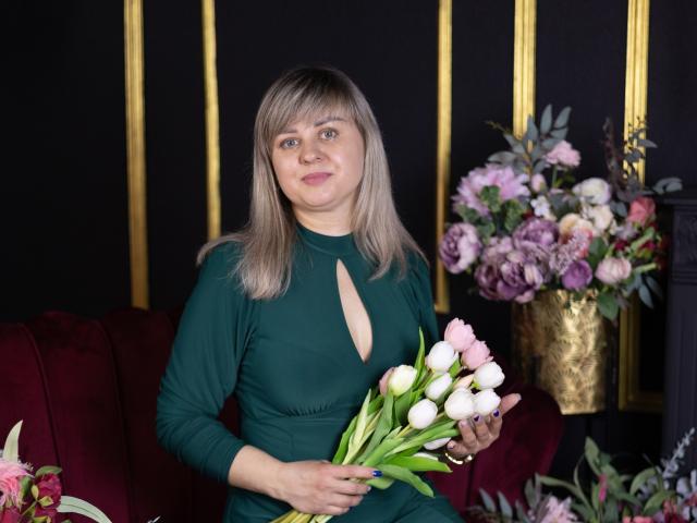 Veronica Svitt, com cabelo castanho liso, vestindo um vestido preto com um decote em formato de chave, segurando um buquê de flores brancas e rosas. tulipas, em frente a um arranjo floral, ambiente escuro.
