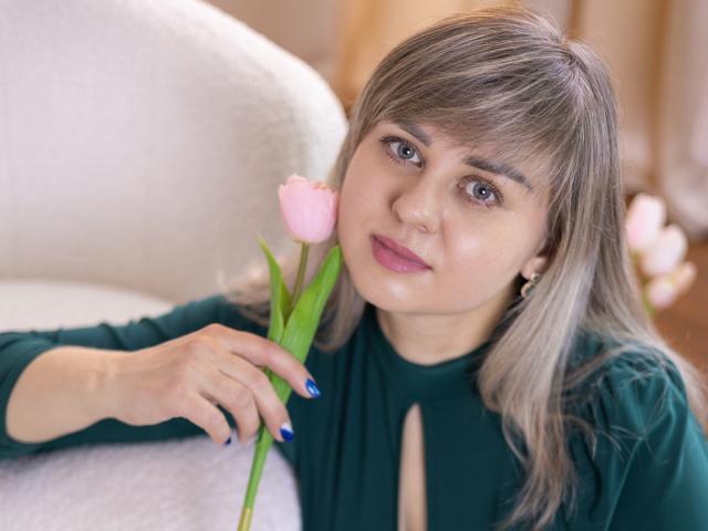 Foto van VeronicaSvitt, een jonge vrouw met lichtbruin haar en een pony, die een roze tulp vasthoudt, gekleed in een zwarte top. direct in de camera kijkend, met een licht verleidelijke uitdrukking.