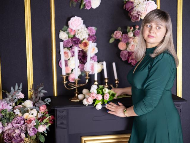 VeronicaSvitt, vestida con un vestido verde, está de pie junto a una consola negra con detalles dorados, rodeada de elementos rosas y arreglos florales blancos y velas. Su expresión es segura, con una ligera sonrisa..