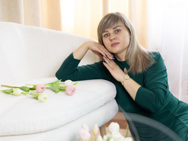 VeronicaSvitt with light brown hair, black long-sleeve top, leaning on white couch, hand on cheek, looking at camera, flowers on couch, soft lighting, gold watch on wrist, seductive gaze.