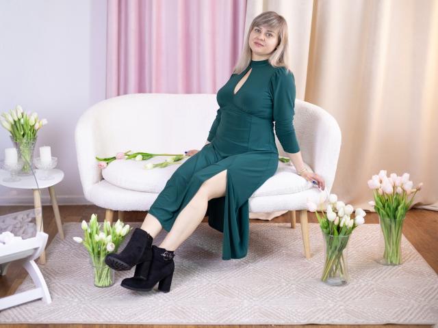 VeronicaSvitt, sentada en un sofá blanco, vestido verde con una abertura alta en la pierna, botas negras hasta el tobillo, piernas separadas, blanco y rosa. Tulipanes alrededor, habitación luminosa, pose informal, tensión sexual, expresión relajada..