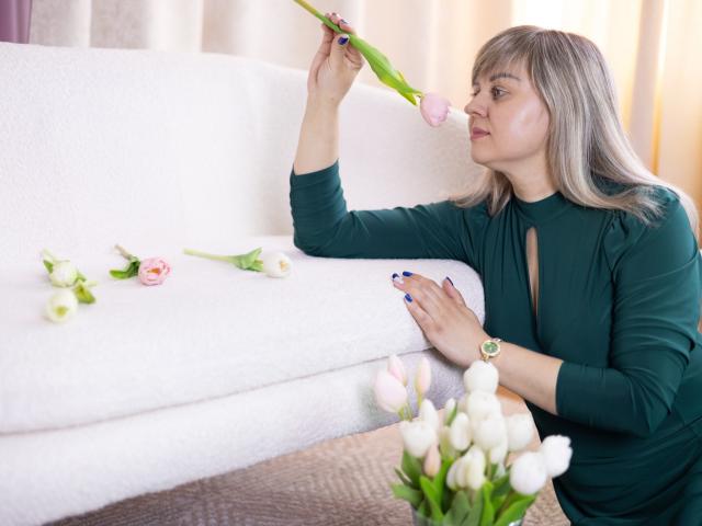 Veronica Svitt, die ein schwarzes Oberteil mit langen Ärmeln trägt, hält eine rosa Tulpe mit ihrer rechten Hand dicht vor ihr Gesicht. Weiße Blumen im Vordergrund, unscharf. Helles, natürliches Licht, lockere Pose..