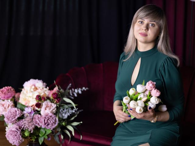 VeronicaSvitt, een blonde vrouw met steil haar, gekleed in een zwarte jurk met een V-hals, houdt een boeket roze bloemen vast. Witte en paarse bloemen. Zwak verlicht, rode achtergrond.
