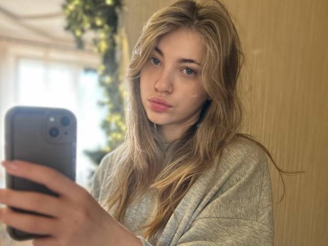 Photographie d'une jeune femme, ShareGullage, avec de longs cheveux blonds ondulés, des lèvres pulpeuses et des yeux bleus, portant un vêtement gris pull, prenant un selfie avec un smartphone. L'arrière-plan est flou. Incroyablement sexy..
