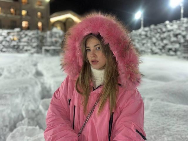Fotografia de uma mulher identificada como ShareGullage, em uma cena noturna com neve. Ela veste um casaco rosa vibrante com um capuz rosa fofo e uma gola alta branca. Uma atmosfera muito fria. O chão coberto de neve..
